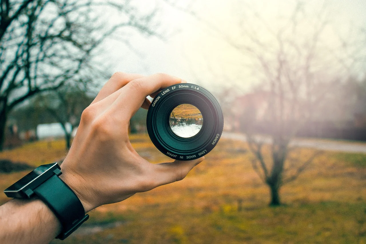 A hand holding a camera lens in front of trees